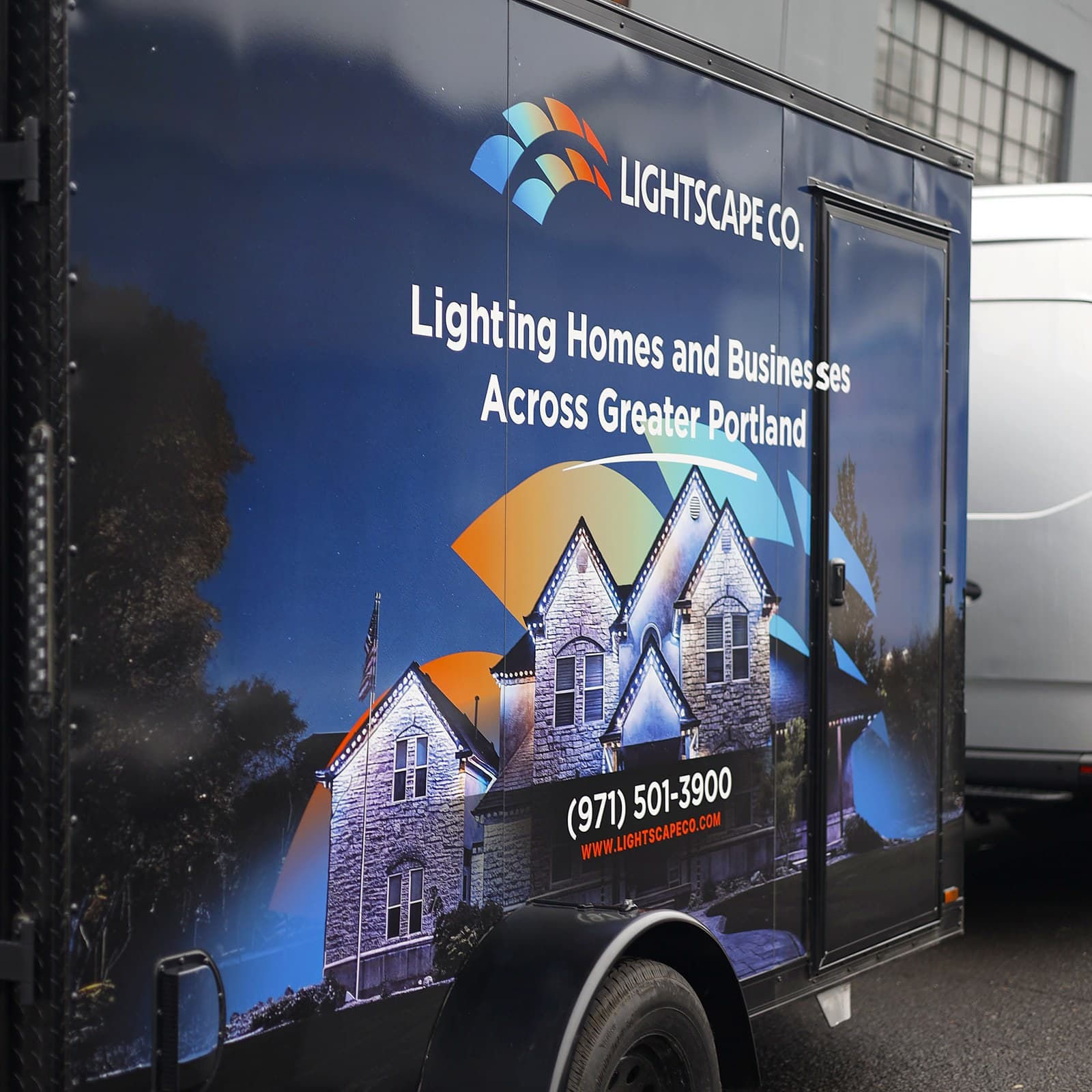 An enclosed trailer wrapped in a printed graphic of a lit house at dusk, seen along its side