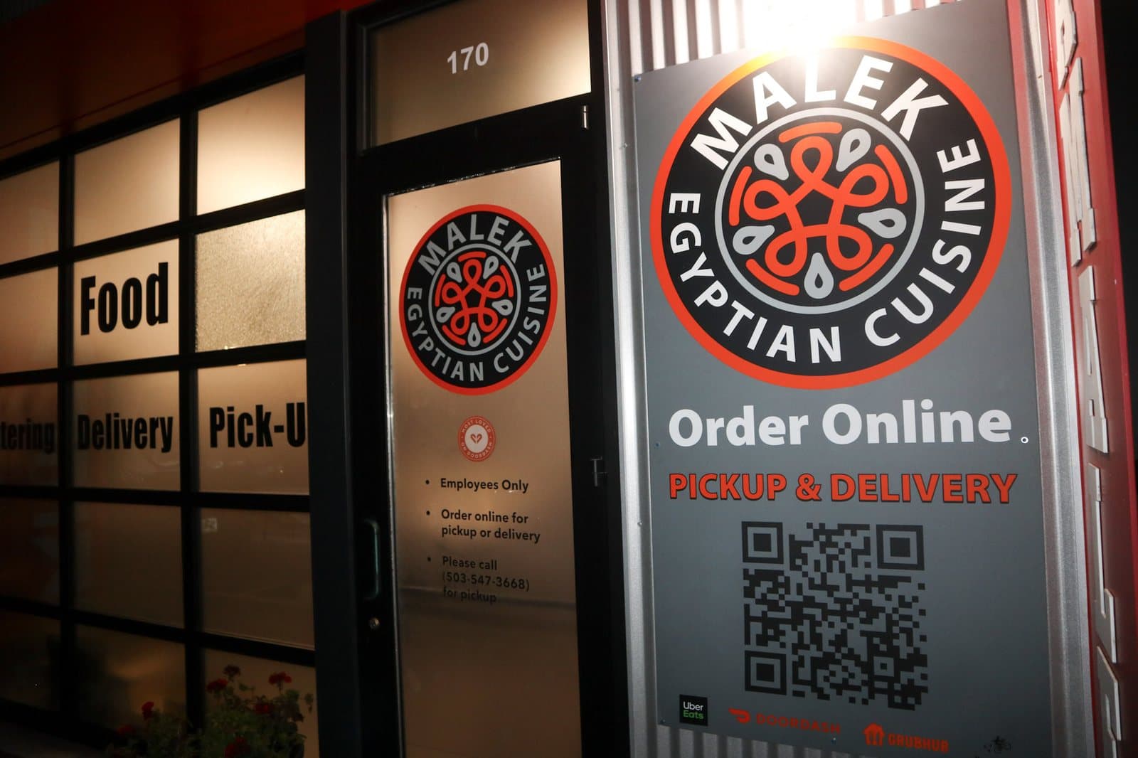 A restaurant front at night: lettering on the windows, a logo on the door and a large printed sign panel