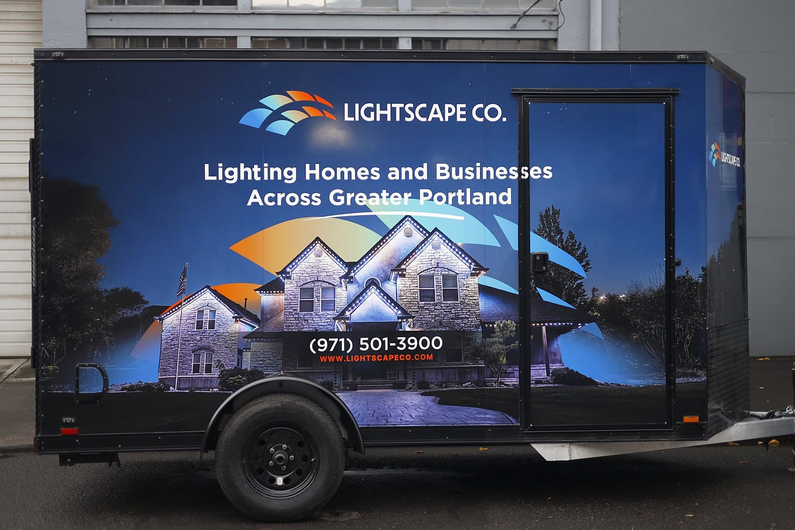 An enclosed trailer wrapped edge to edge in a printed graphic of a lit house at dusk, seen from the side